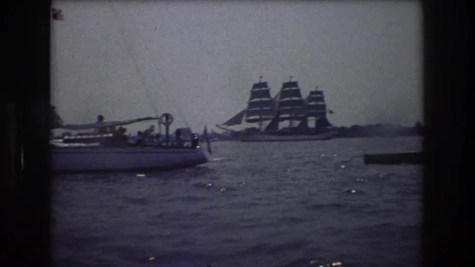 1976 8mm frame on New York City waters showing a small passenger boat with people aboard and a large multi-masted sailing ship farther across the harbor.