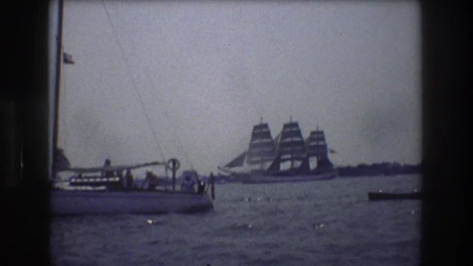 1976 New York City harbor frame showing a small boat with passengers at left, a large multi-masted sailing ship in the distance, and sunlit water under a pale sky.