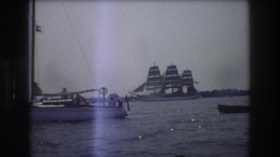 1976 New York City 8mm frame showing a small sailboat with people aboard and a distant three-masted sailing ship under sail across the water.