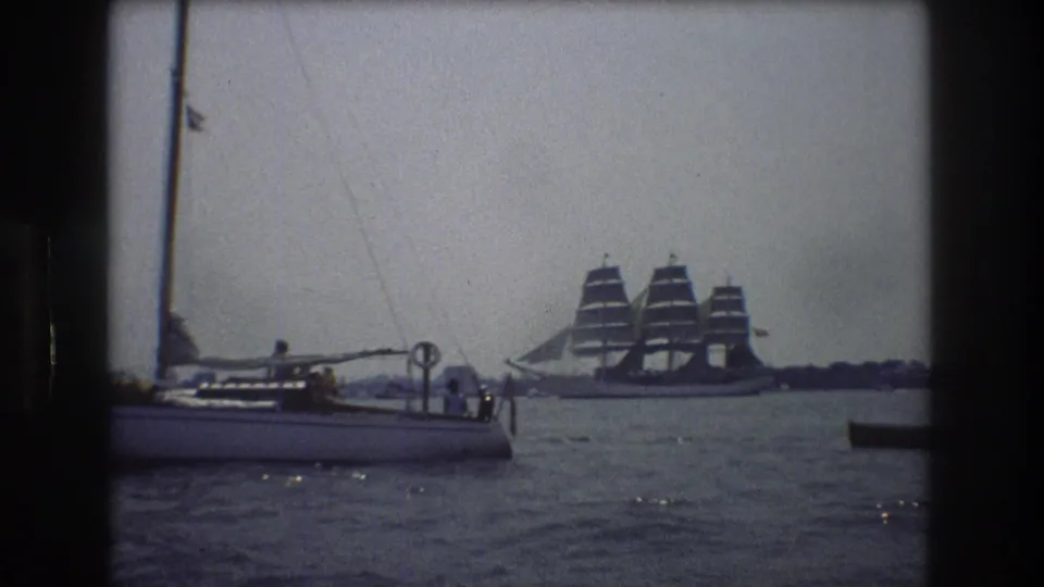 1976 New York City home-movie frame with a foreground sailboat, small figures aboard, and a large three-masted sailing ship beyond it on calm harbor water.