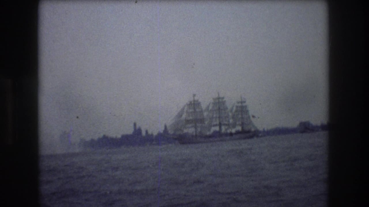 1976 New York City home-movie frame showing a large multi-masted sailing ship moving across calm harbor water with a faint distant shoreline.