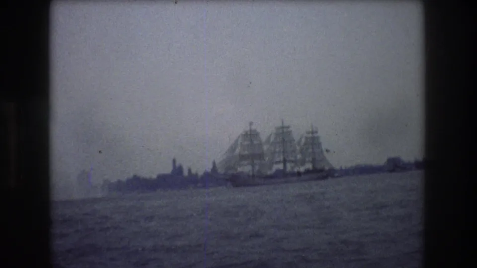 1976 New York City home-movie frame showing a large multi-masted sailing ship moving across calm harbor water with a faint distant shoreline.