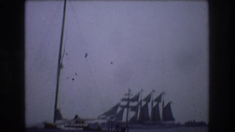 1976 New York City home-movie frame showing a foreground sailboat mast and hull with a large multi-masted sailing ship farther out on calm water.
