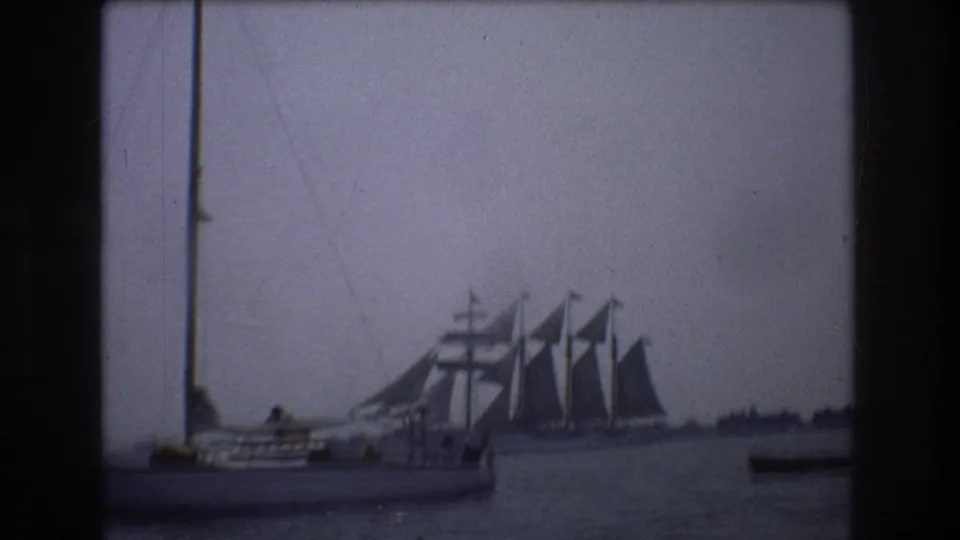 1976 New York City home-movie frame showing a small foreground sailboat with indistinct people aboard and a large multi-masted sailing ship behind it on harbor water.