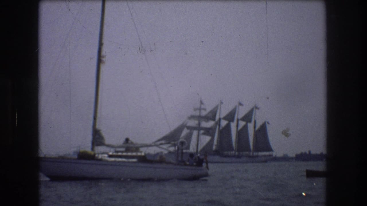 1976 New York City 8mm frame showing a small sailboat passing in front of a larger multi-masted sailing vessel on calm harbor water.