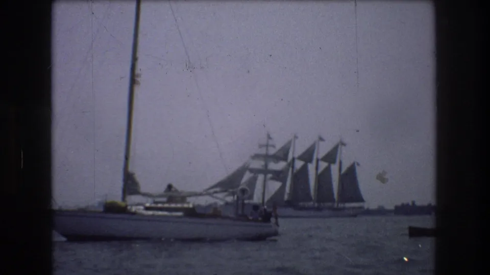 1976 New York City 8mm frame showing a small sailboat passing in front of a larger multi-masted sailing vessel on calm harbor water.