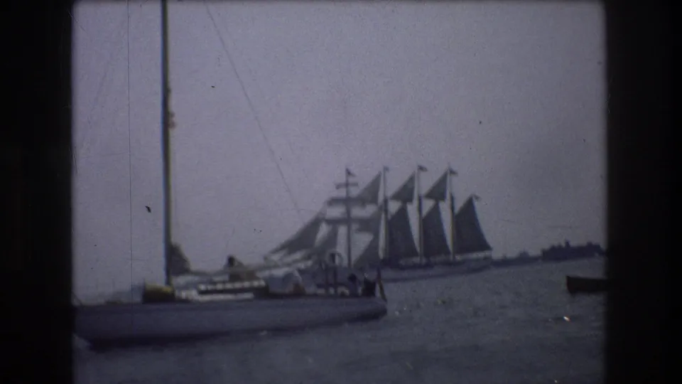 1976 New York City home-movie frame with a sailboat in the foreground and a larger sailing vessel beyond it under a gray sky.