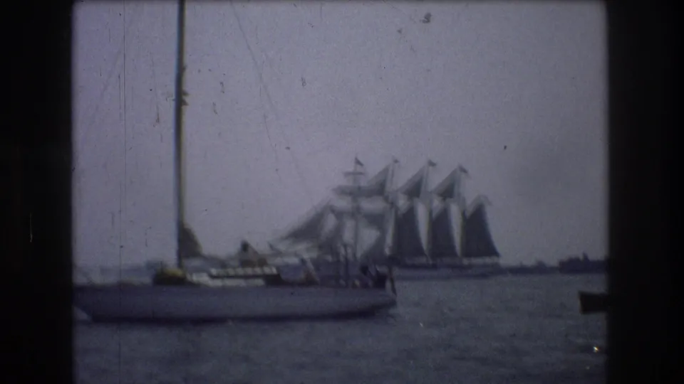 Soft 8mm frame from 1976 New York City showing a foreground sailboat, visible passengers, and a larger sailing vessel in the background.