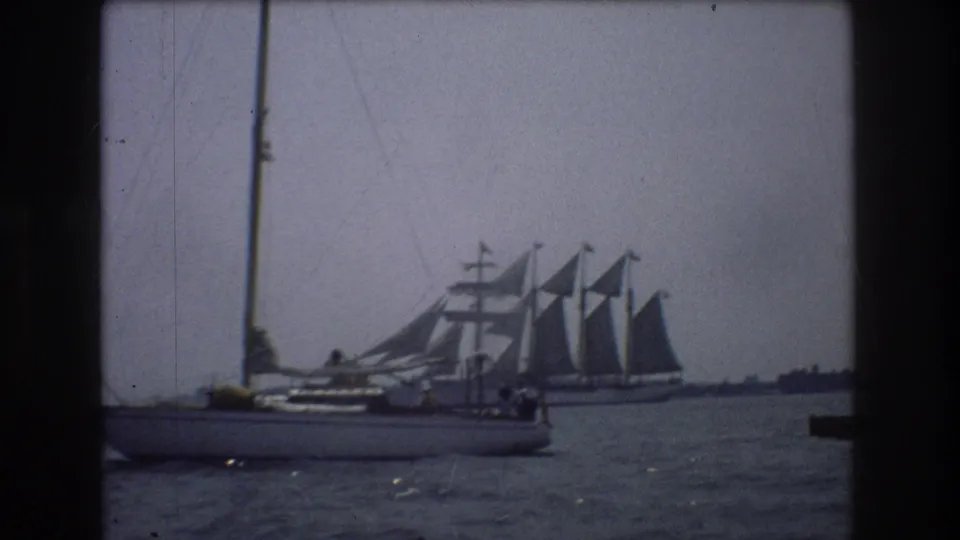 1976 New York City 8mm frame showing the foreground sailboat slightly farther along, with the larger multi-masted vessel still visible behind it.