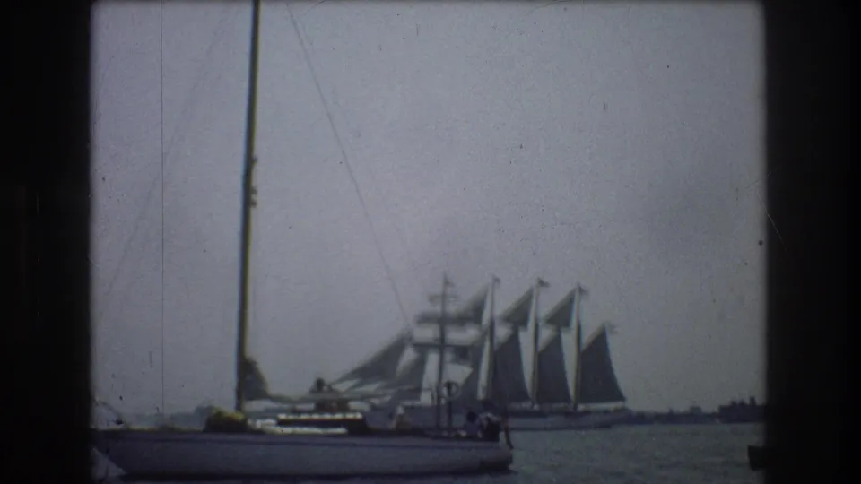 1976 New York City-area harbor footage showing a foreground sailboat with a tall mast and a larger multi-masted sailing vessel under sail in the background.