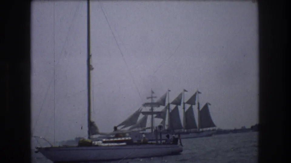 A small sailboat with crew-sized figures sits in front of a larger multi-masted sailing ship on gray-blue water near New York City in the supplied 1976 home-movie metadata.