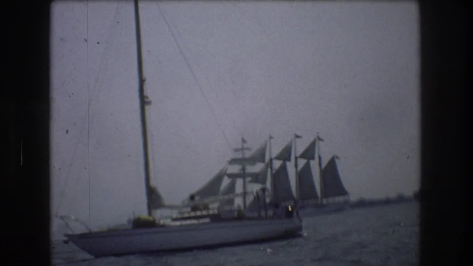 Soft 8mm frame of a foreground sailboat crossing the lower harbor view, with a larger multi-masted vessel under sail behind it in the New York City-area setting supplied by metadata.