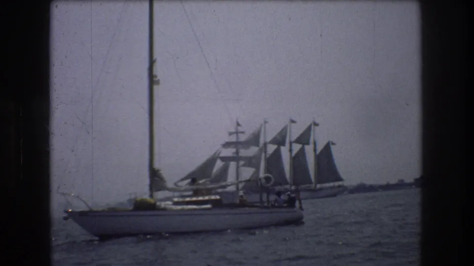 1976 home-movie still from the New York City area showing a small sailboat with visible rigging in front of a larger multi-masted sailing ship under muted overcast light.