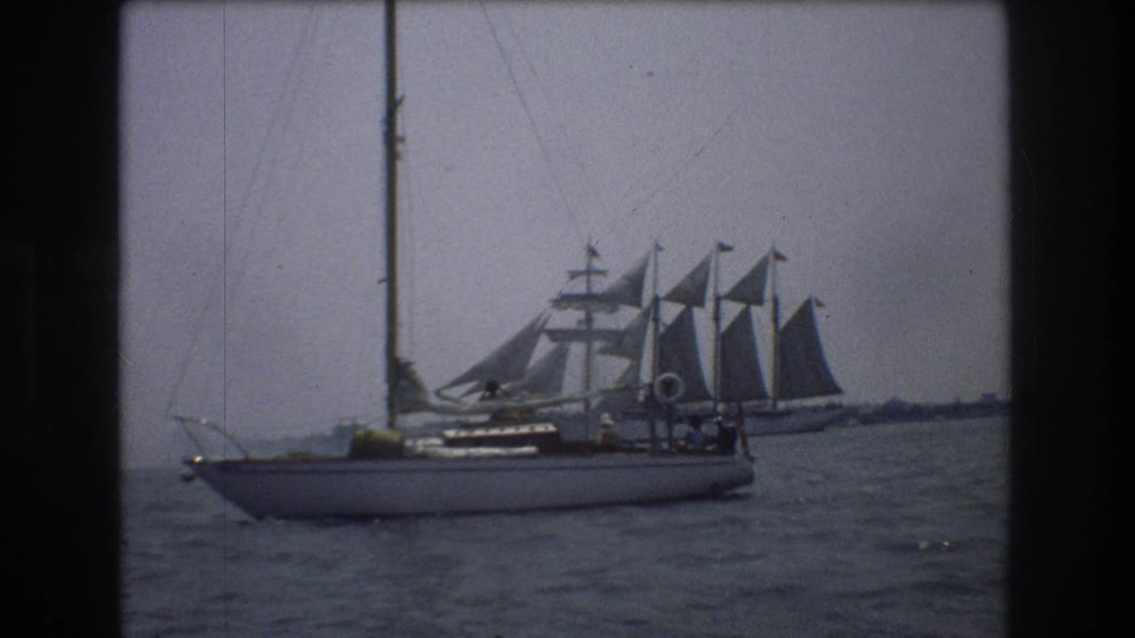 1976 New York City home-movie frame showing a sailboat on harbor water with a larger multi-sail vessel in the background.
