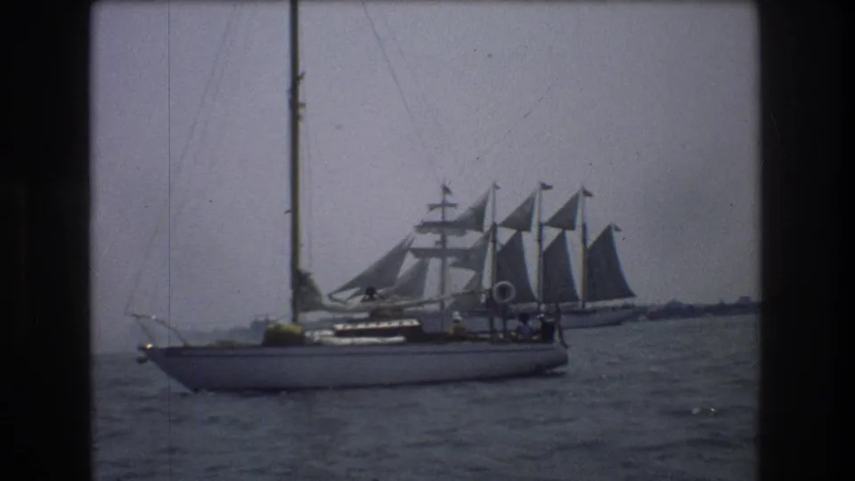 1976 New York City home-movie frame showing a sailboat on harbor water with a larger multi-sail vessel in the background.
