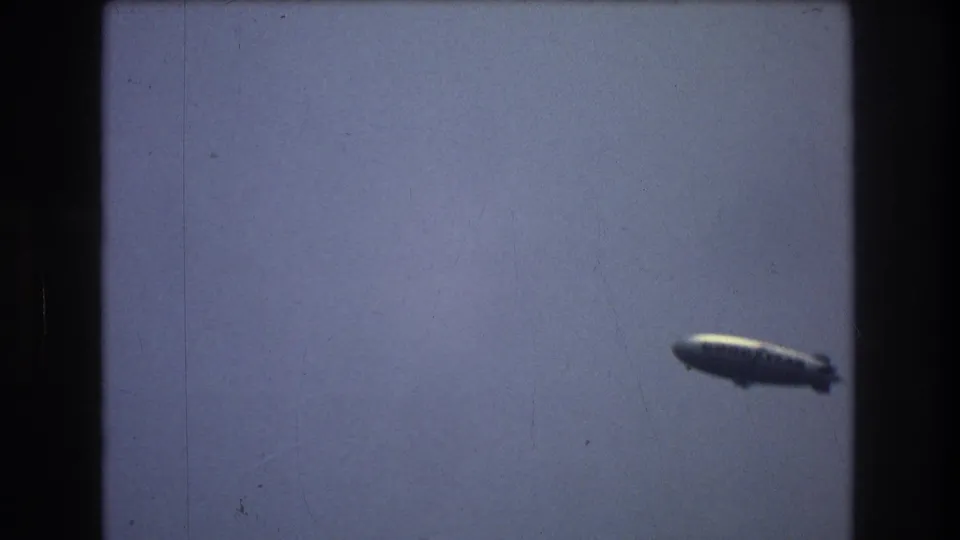 Distant airship or blimp crossing a blue-gray sky in 1976 New York City home-movie footage.