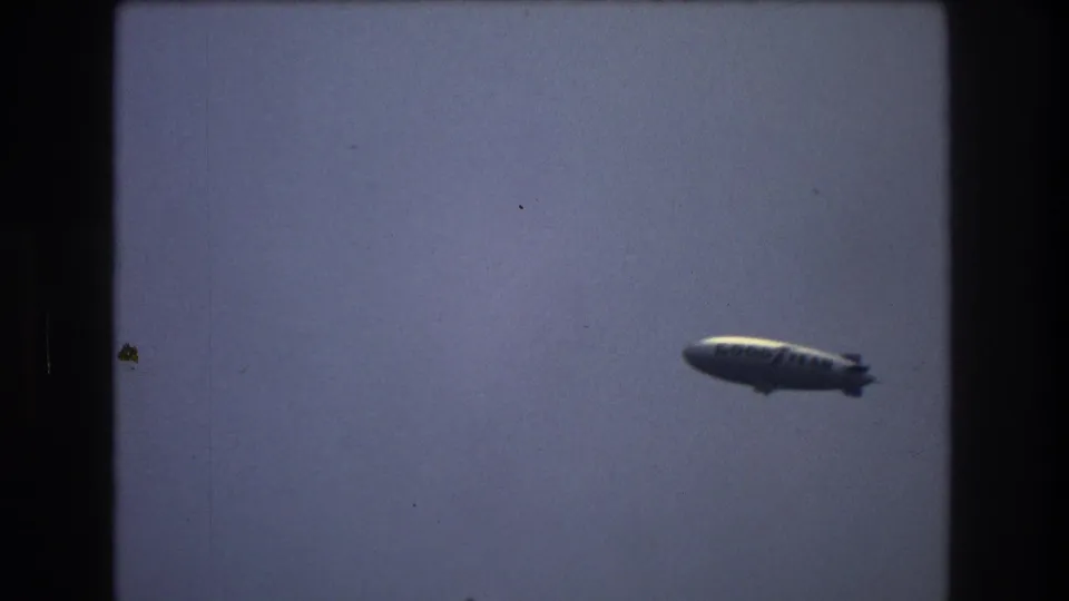 Soft 8mm frame of a dark airship shape passing through open sky, with no visible ground reference.