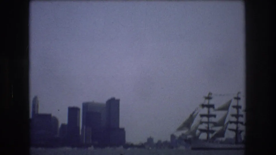 A tall ship with sails and rigging passes across New York City harbor with the skyline silhouetted in the background.