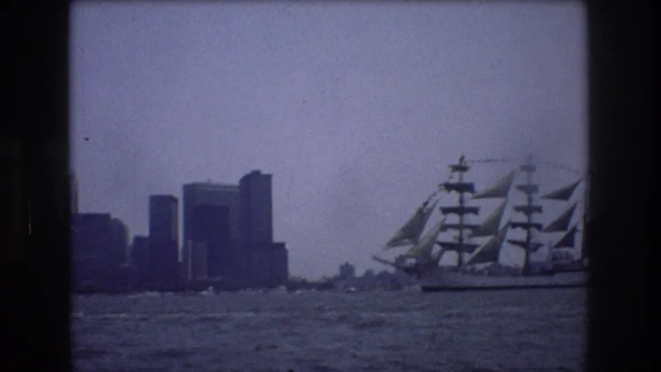 1976 New York City waterfront view with a multi-masted sailing ship moving across choppy water before a distant skyline.