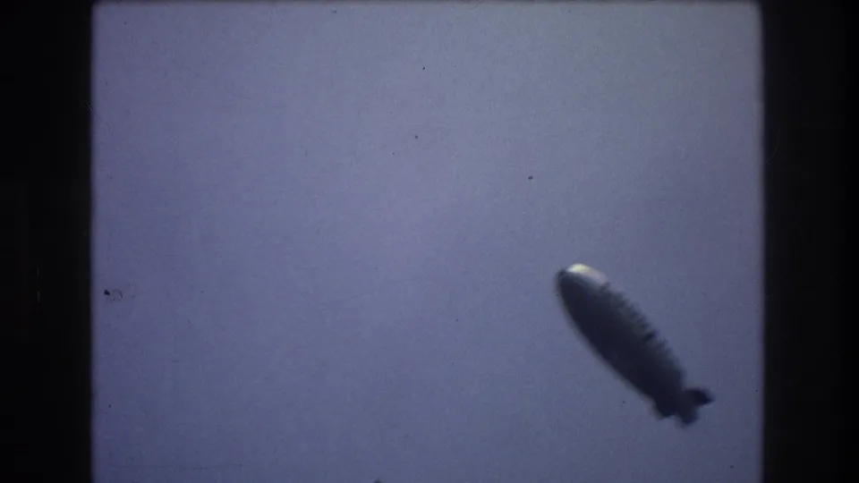 A silver blimp appears lower in the frame, angled through the hazy sky in the New York City clip.