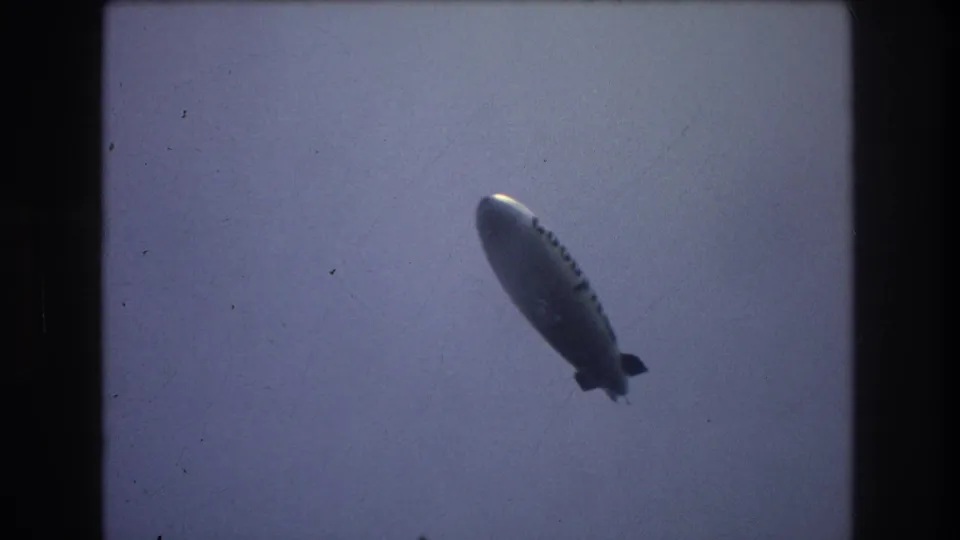 Silver blimp continues across the hazy sky in the final frame of the 1976 New York City home movie clip.