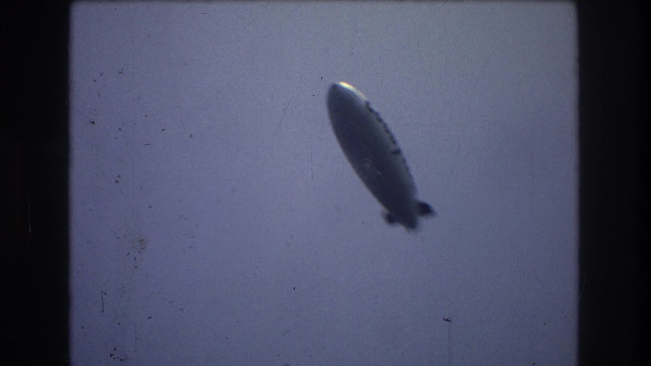 A Goodyear airship appears high against a pale blue-gray sky in 1970s 8mm New York City footage, with soft focus and dark film-frame edges.