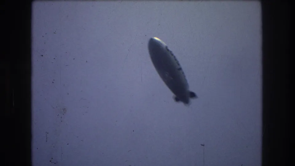 A Goodyear airship appears high against a pale blue-gray sky in 1970s 8mm New York City footage, with soft focus and dark film-frame edges.