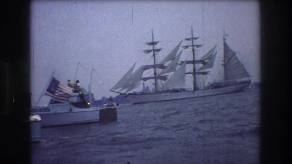 A small boat carrying people and an American flag sits on the water near a large multi-masted sailing ship, in soft 1976 New York City 8mm harbor footage.
