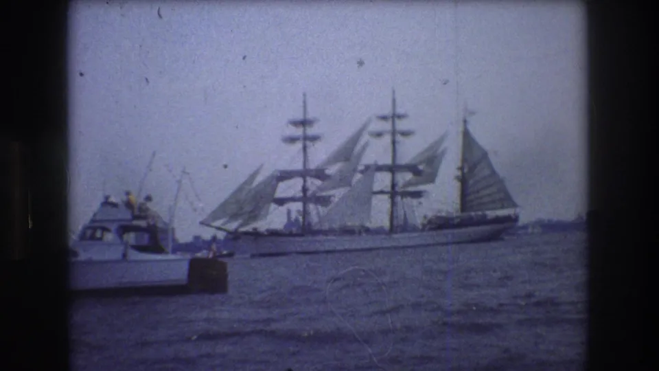 1976 New York City home-movie frame showing a multi-masted sailing ship with sails raised passing behind a small motorboat on harbor water.