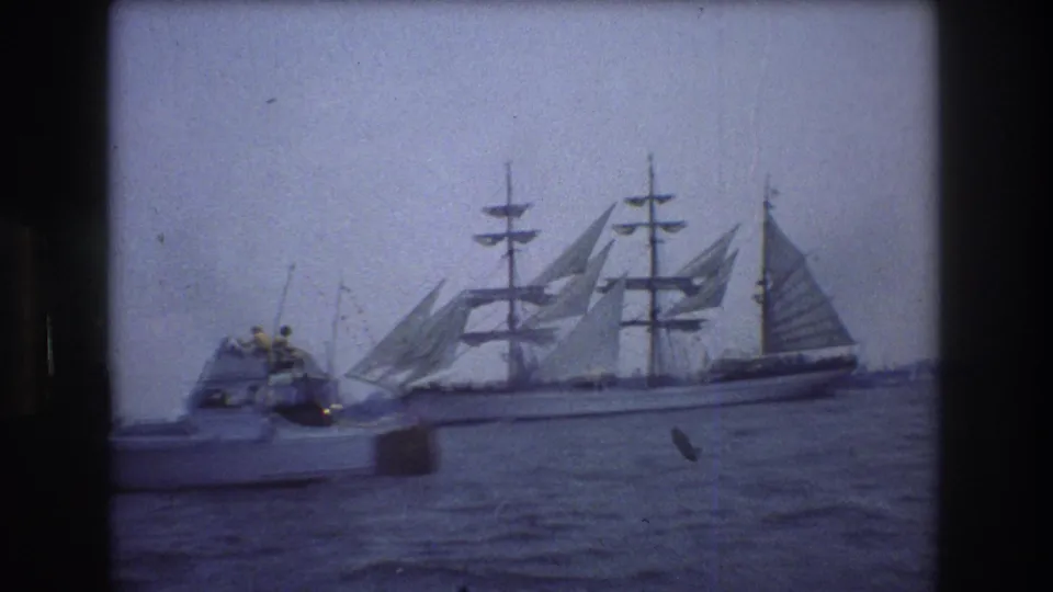 1976 New York City harbor footage frame with a small motorboat in front of a large multi-masted sailing ship under overcast light.