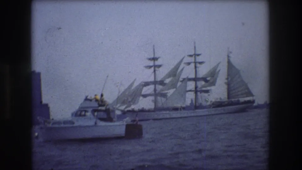 A 1976 New York City 8mm frame shows a multi-masted sailing ship beyond a small motorboat, with soft focus, blue tint, and dark frame edges.