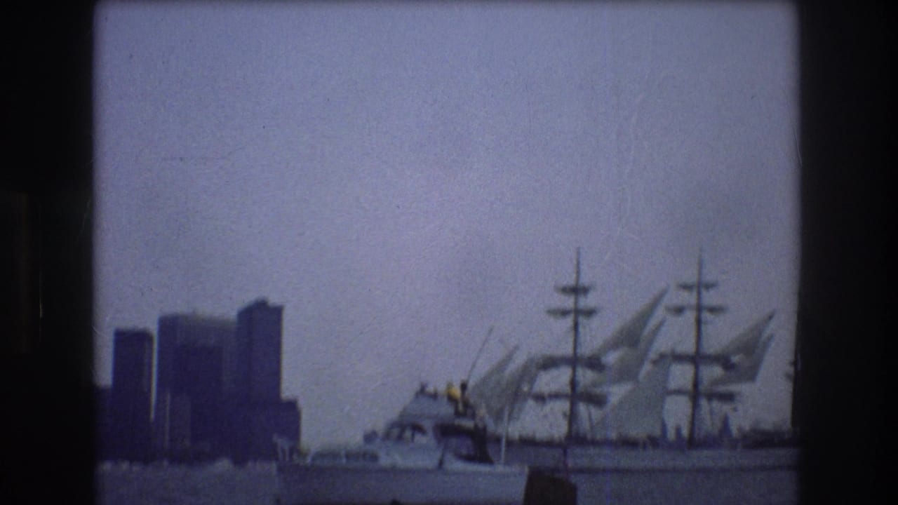 Three-masted sailing ship and small motorboat on harbor water with city towers in the background.