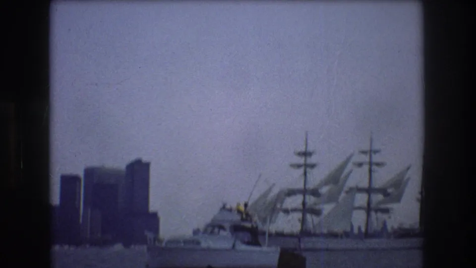 1976 New York City harbor view with a three-masted sailing ship passing behind a smaller motorboat and dark skyline towers under an overcast sky.
