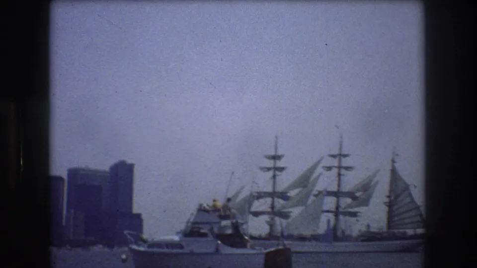 The smaller motorboat sits in the foreground as the three-masted sailing ship moves across New York Harbor with skyline towers behind it.