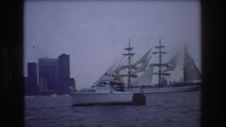 A small motorboat passes across the foreground while the three-masted sailing ship remains behind it on 1976 New York City harbor water.