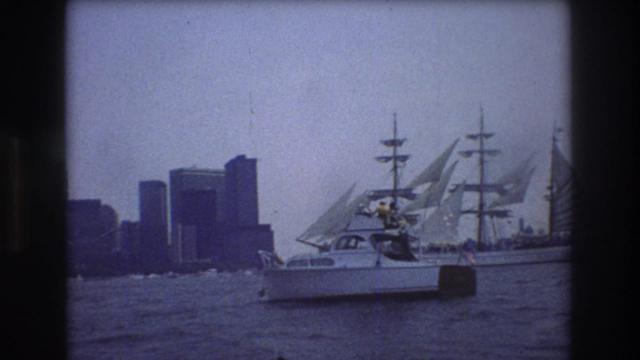 1976 8mm home-movie frame of a small motorboat crossing harbor water in front of a multi-masted sailing vessel and hazy Manhattan high-rises.