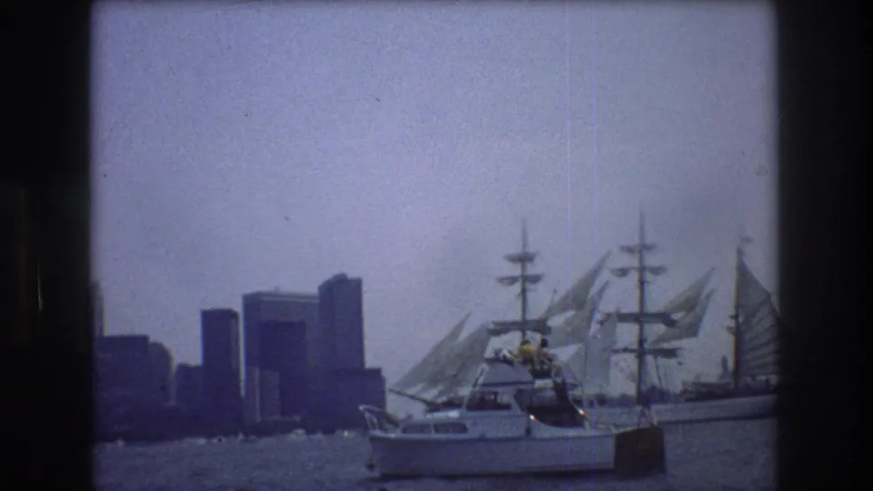 A small motorboat sits in the foreground while a large sailboat and hazy New York City skyline fill the background in this 1976 harbor view.