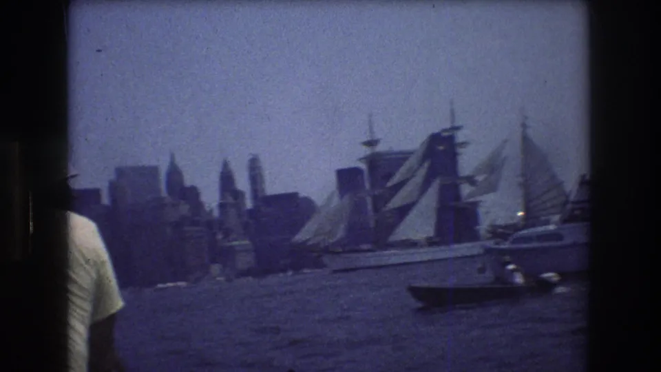 Handheld 8mm frame showing a sailboat near the Manhattan skyline with a small dark boat in front and part of a person entering the left edge.