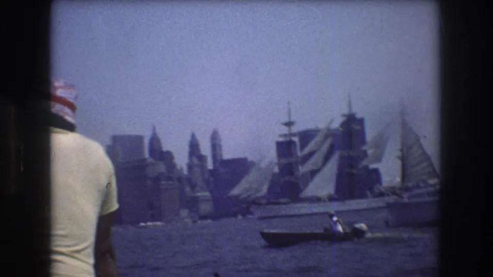 A person in a light shirt and patterned hat is partially visible in the foreground as sailboats and a small boat move across New York harbor in 1976.