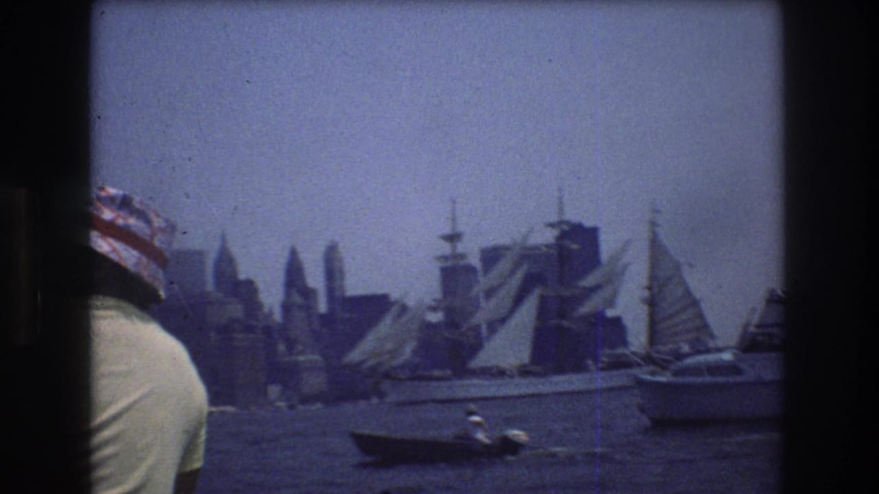 1976 New York City harbor view with a man in a striped hat at the edge of frame, sailboats on the water, and the Manhattan skyline in haze.