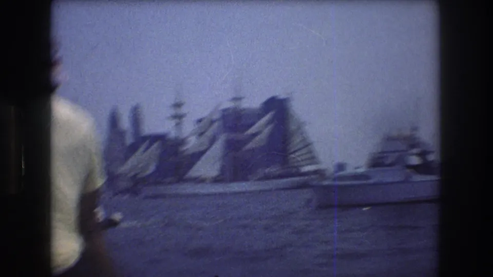 Motion-blurred 8mm view of New York Harbor with sailboats and skyline, partly blocked by a foreground figure at the left edge.
