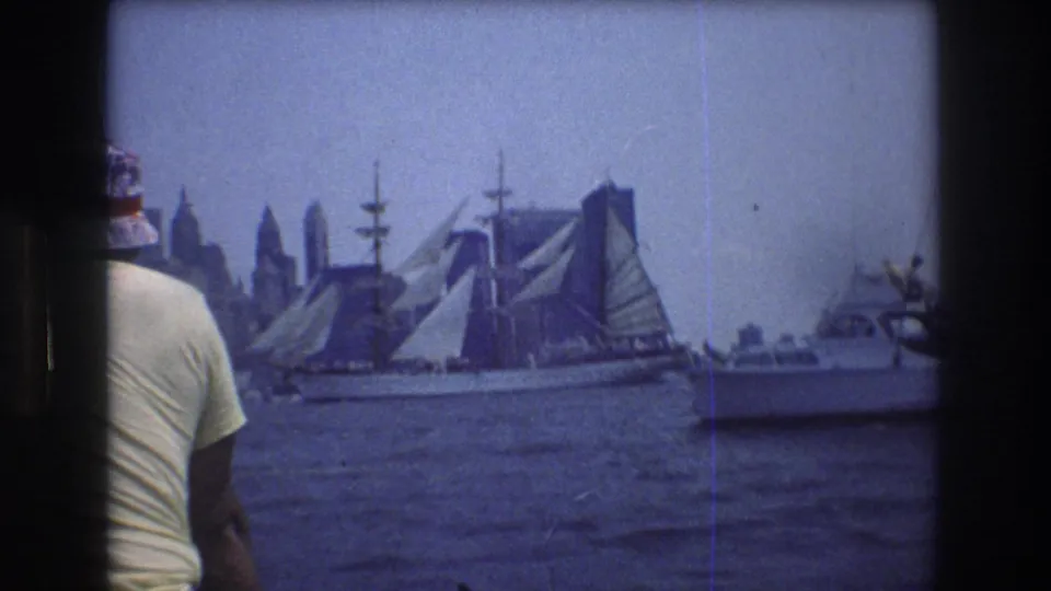 1976 New York Harbor frame showing a large sailing ship with raised sails, smaller craft, a hazy Manhattan skyline, and a foreground man at left.