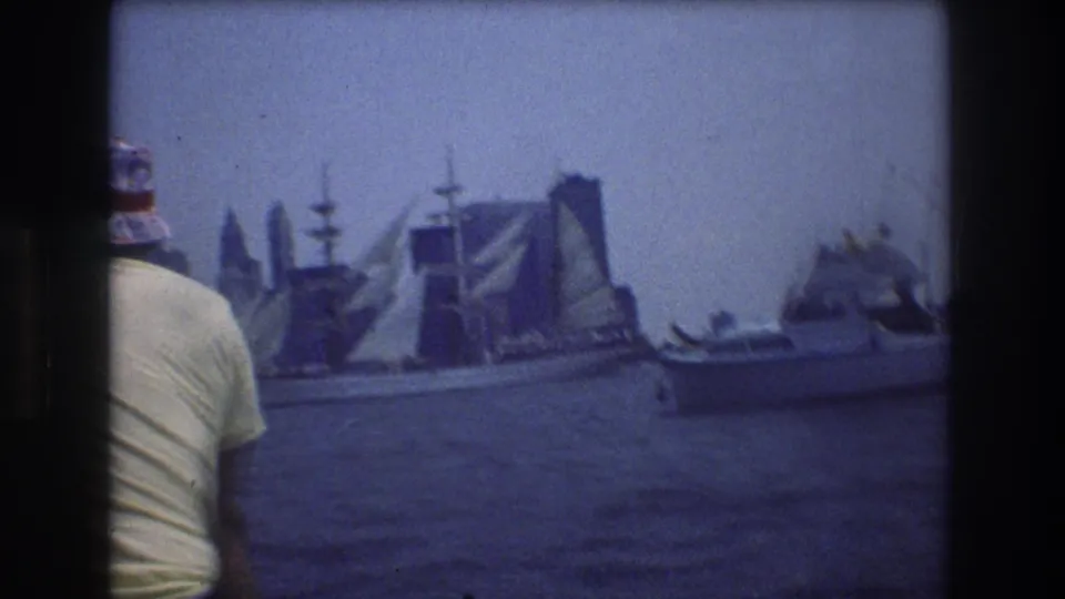 1976 8mm New York Harbor view with a foreground passenger, a multi-masted sailing ship, smaller boats, and the hazy Manhattan waterfront in the distance.