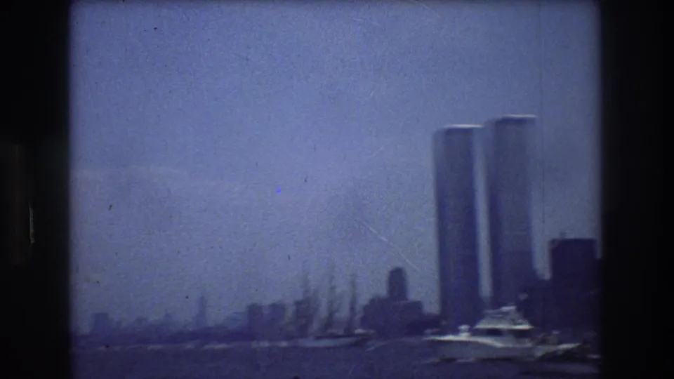 Soft, blue-tinted 1976 8mm frame showing the Twin Towers above the Manhattan waterfront, with masts or rigging near the shoreline and harbor water in front.