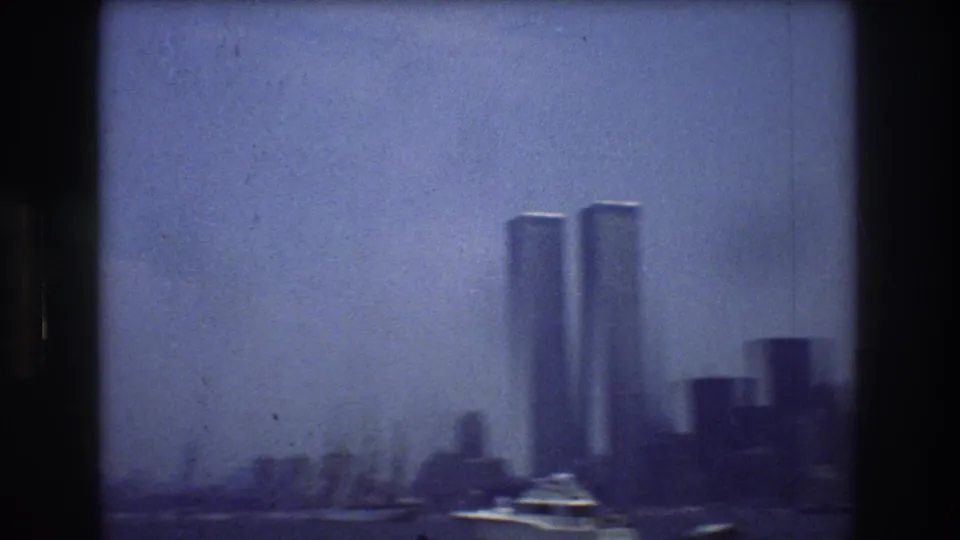 1976 8mm harbor view with the Twin Towers at center right, dark waterfront buildings, a white motorboat, and soft haze over Manhattan.