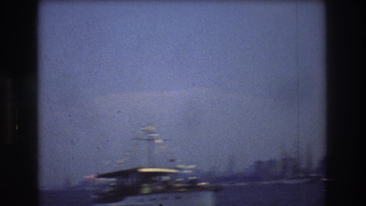 1976 New York City harbor view with a large covered-deck boat moving across hazy water and a soft distant skyline in the background.