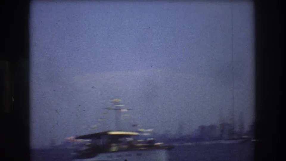 1976 New York City harbor view with a large covered-deck boat moving across hazy water and a soft distant skyline in the background.