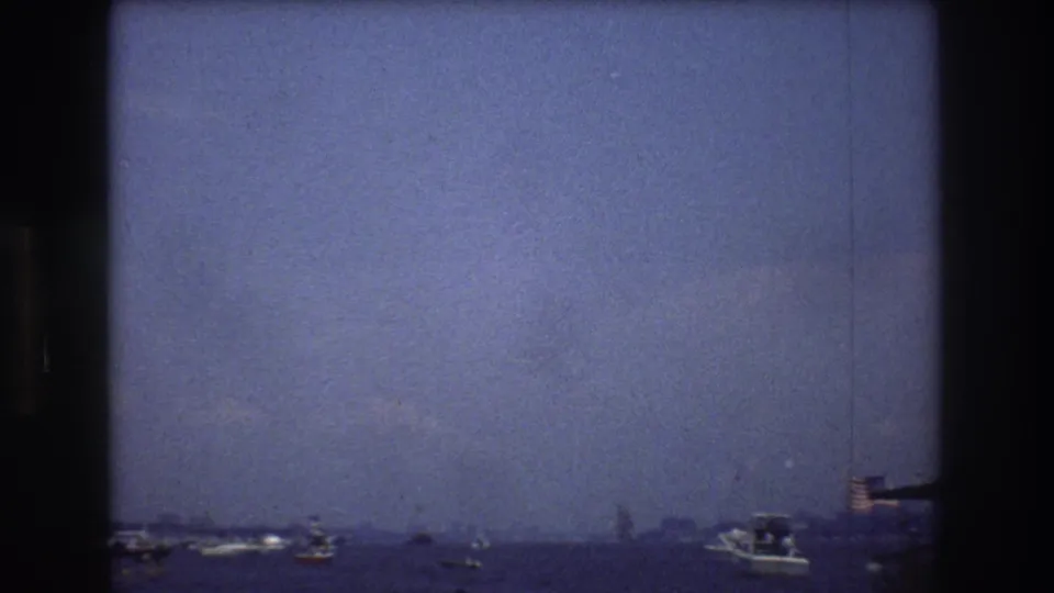 1976 New York City harbor view with scattered small boats, hazy distance, and a small American flag visible near the lower right edge.