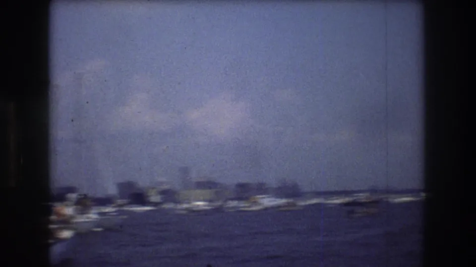1976 New York City harbor shot with distant low skyline forms, boats near the waterline, and soft clouds across a blue-tinted sky.