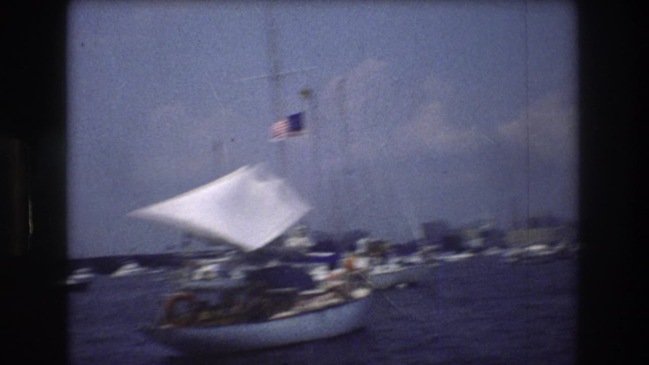 1976 New York City harbor home-movie frame showing a small sailboat with a white sail and American flag moving past other boats on blue water.